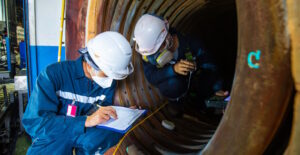 Two workers in hard hats and masks in an industrial working environment with an attendant sheet and clip board.